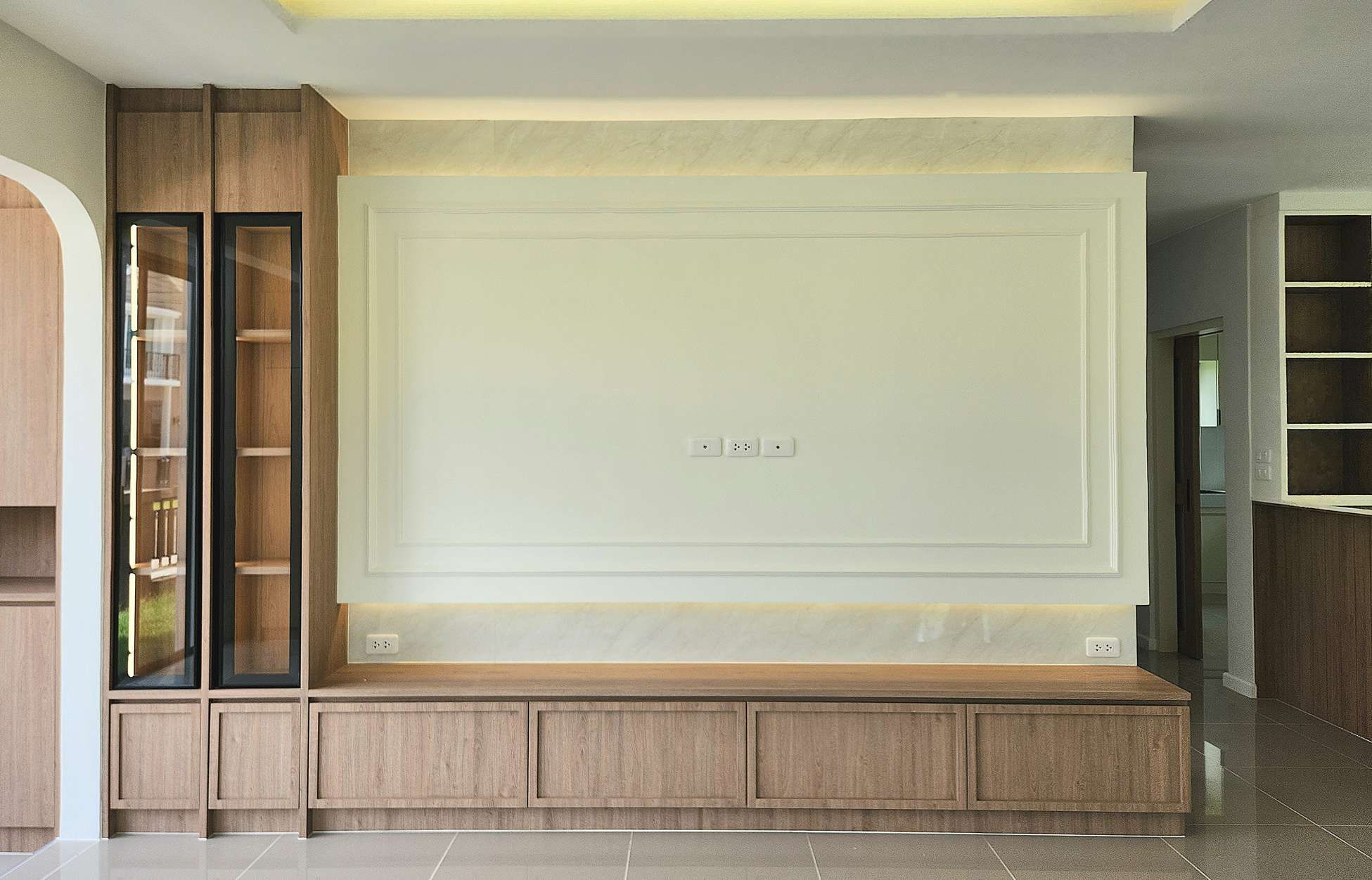 A living room feature wall with a framed white panel, cove lighting, a built-in oak cabinet and lit glass display cases, built by OUT at Laddarom Tiwanon